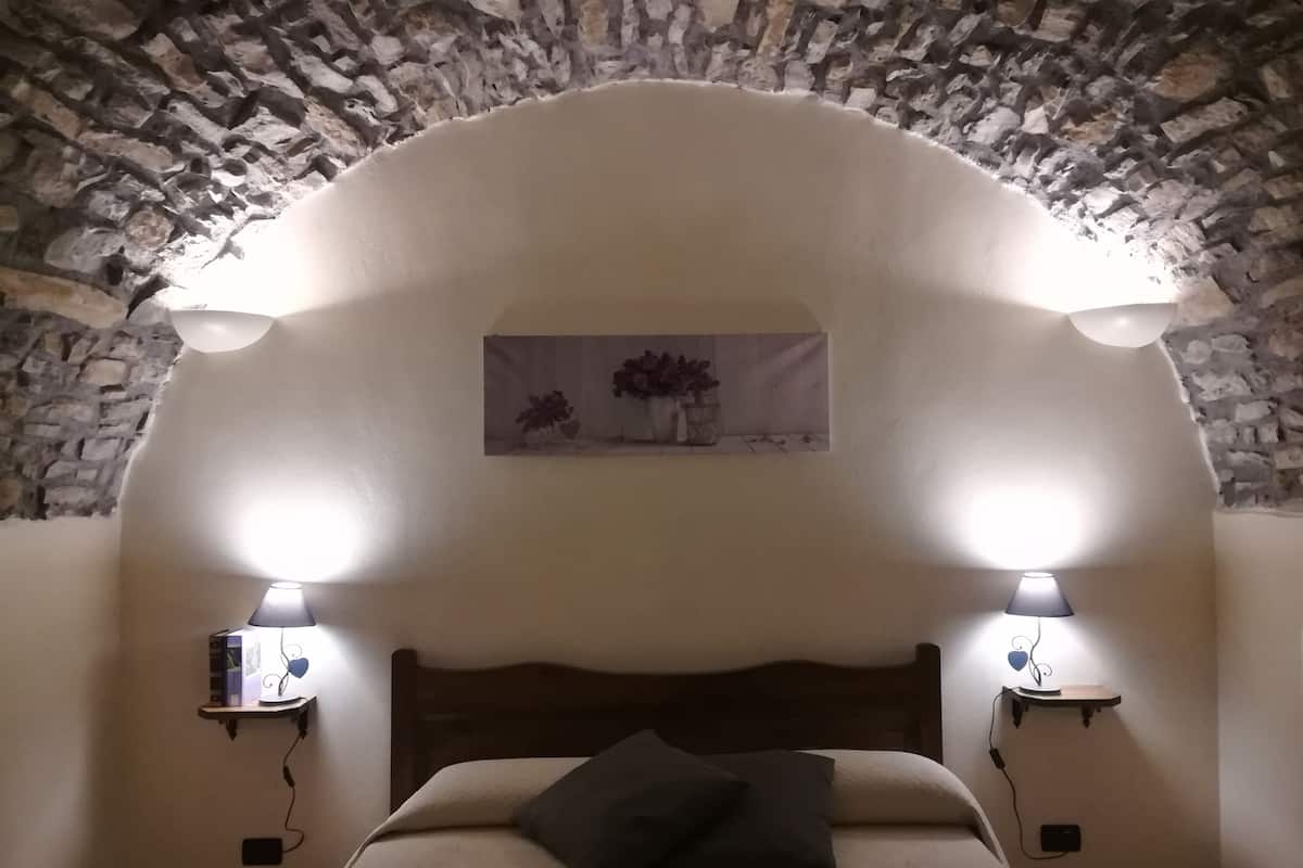 A bedroom with a stone wall, a wooden headboard, two bedside lamps, and a painting above the bed.