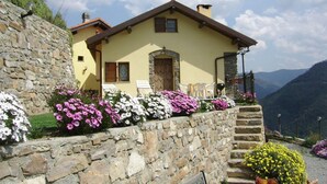 A stone-walled building with a yellow facade, a wooden door, and a small porch with white chairs. - Agriturismo Al Pagan (Pigna)