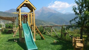 A wooden play structure with a slide, set against a backdrop of mountains and greenery. - Agriturismo Al Pagan (Pigna)