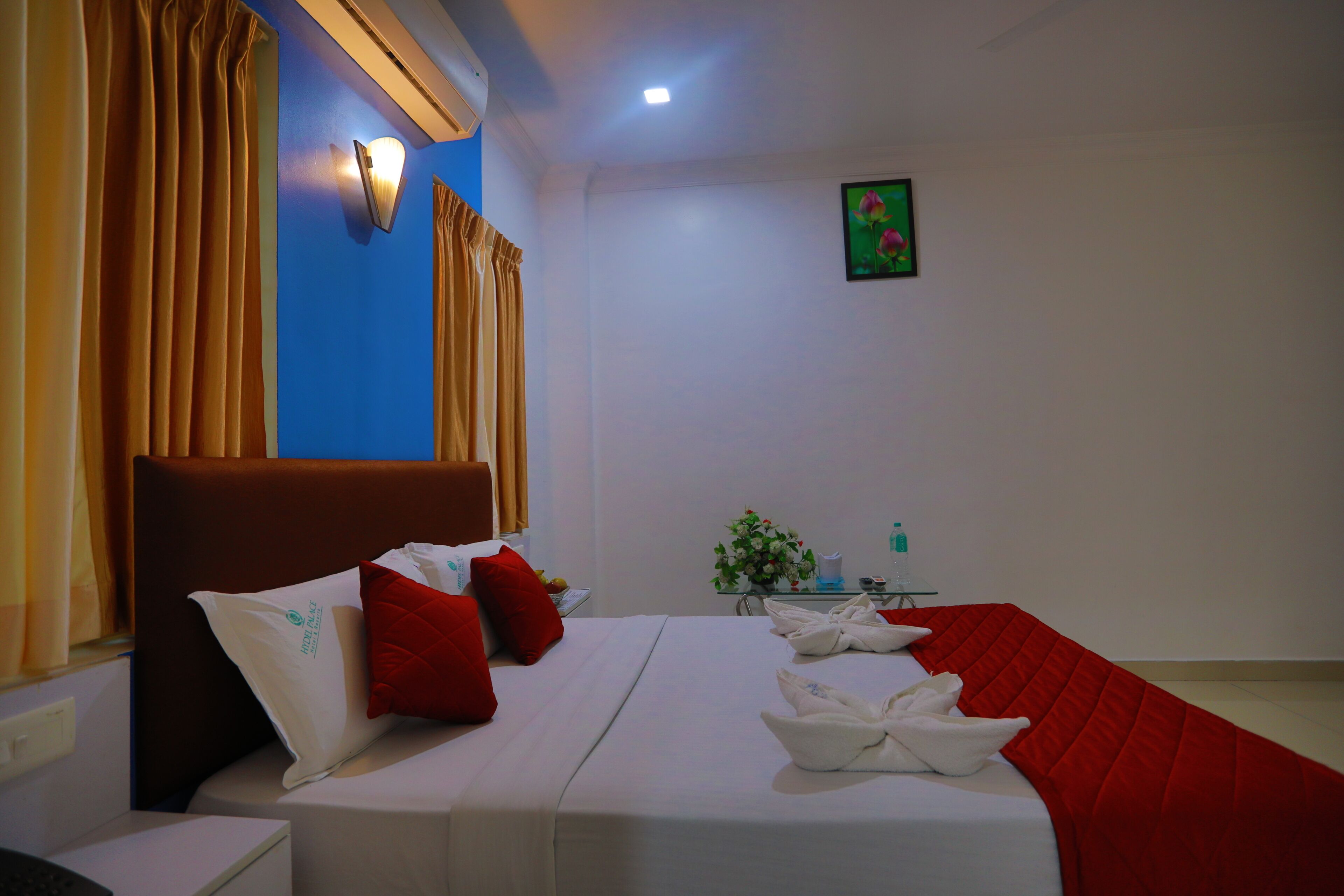 A hotel room with a bed, red and white pillows, a bedside table, a small table with a vase of flowers, and a wall-mounted light fixture.