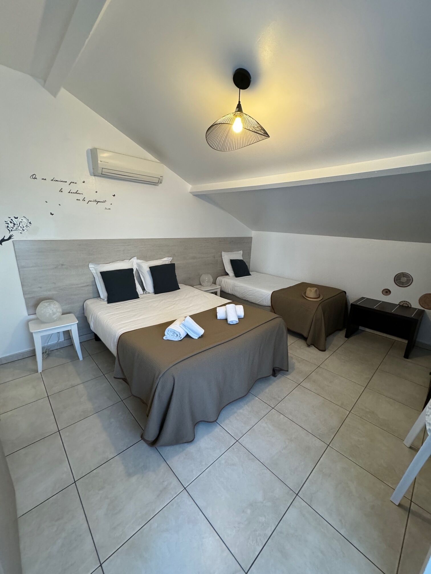 A modern hotel room with two beds, a small table, a bedside table, a wall-mounted air conditioner, and a ceiling light fixture.
