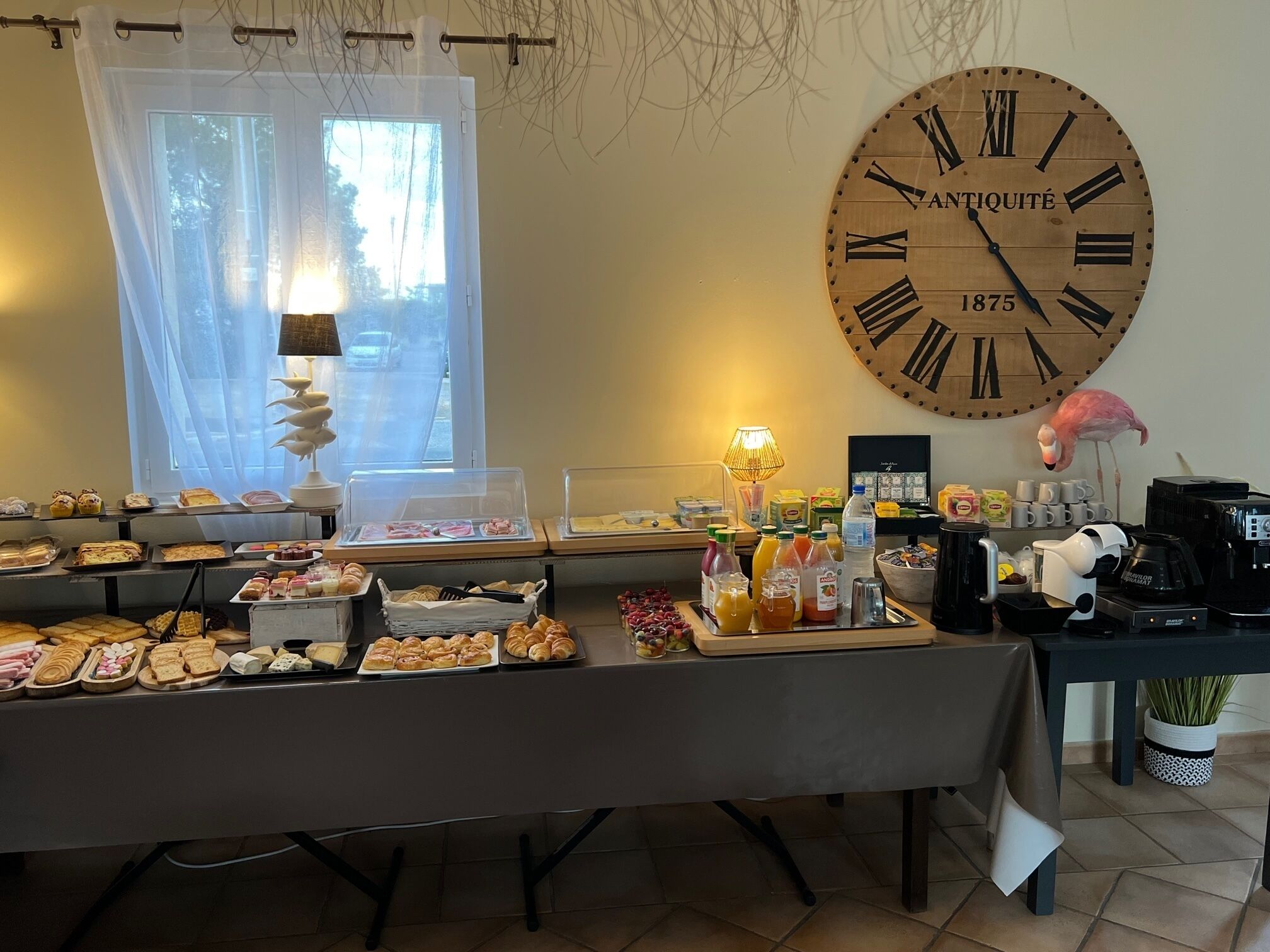 Ein Frühstücksbuffet mit verschiedenen Backwaren und Getränken, eine große antike Uhr und ein dekorativer Flamingo.