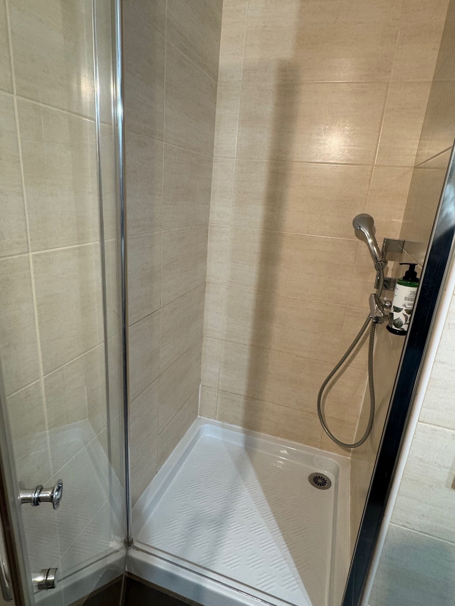 A glass-enclosed shower area with beige tiles, a handheld showerhead, and a bottle of shampoo.