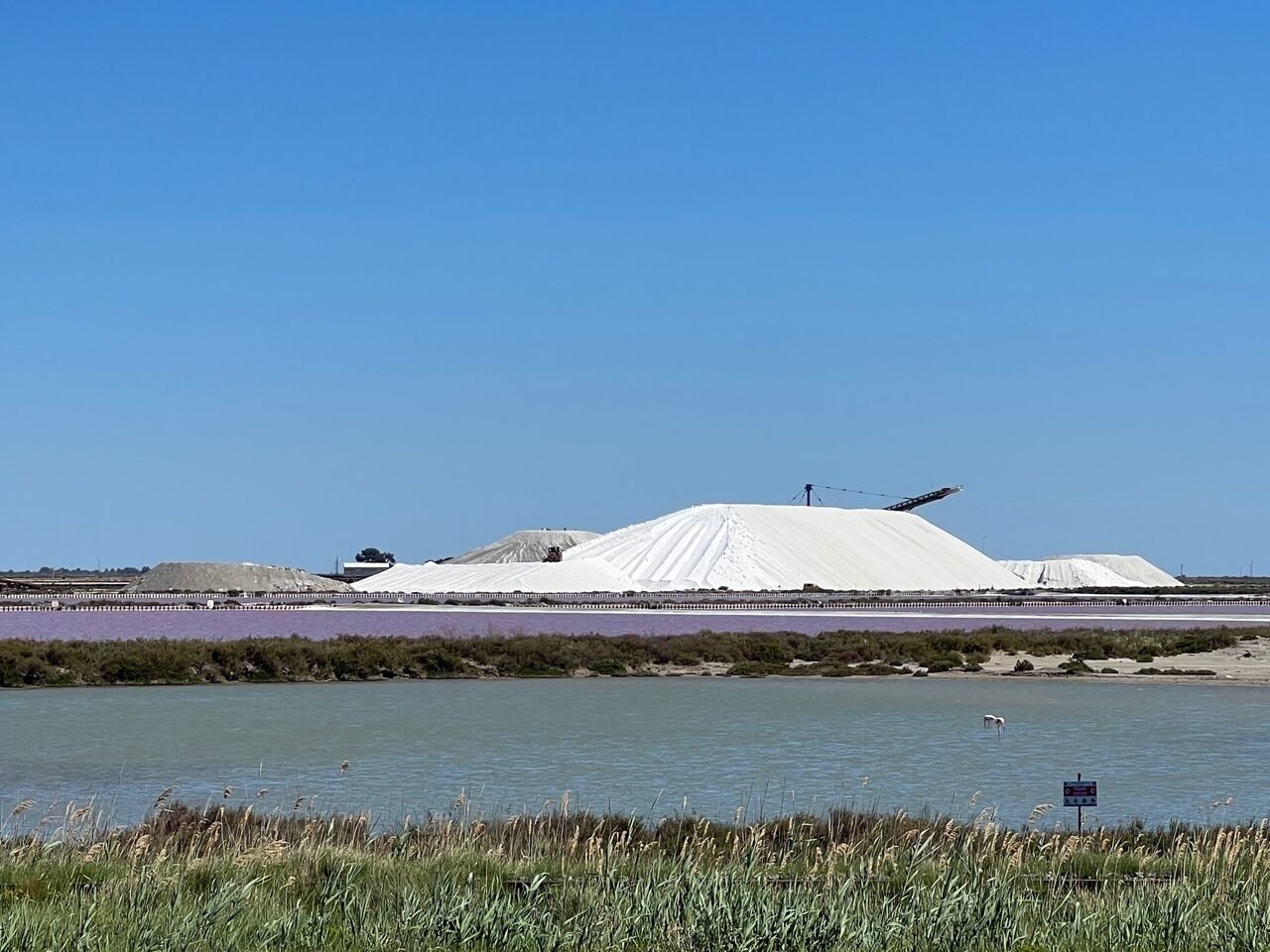 Une grande construction blanche située près d’une étendue d’eau, entourée d’une clôture et de végétation.