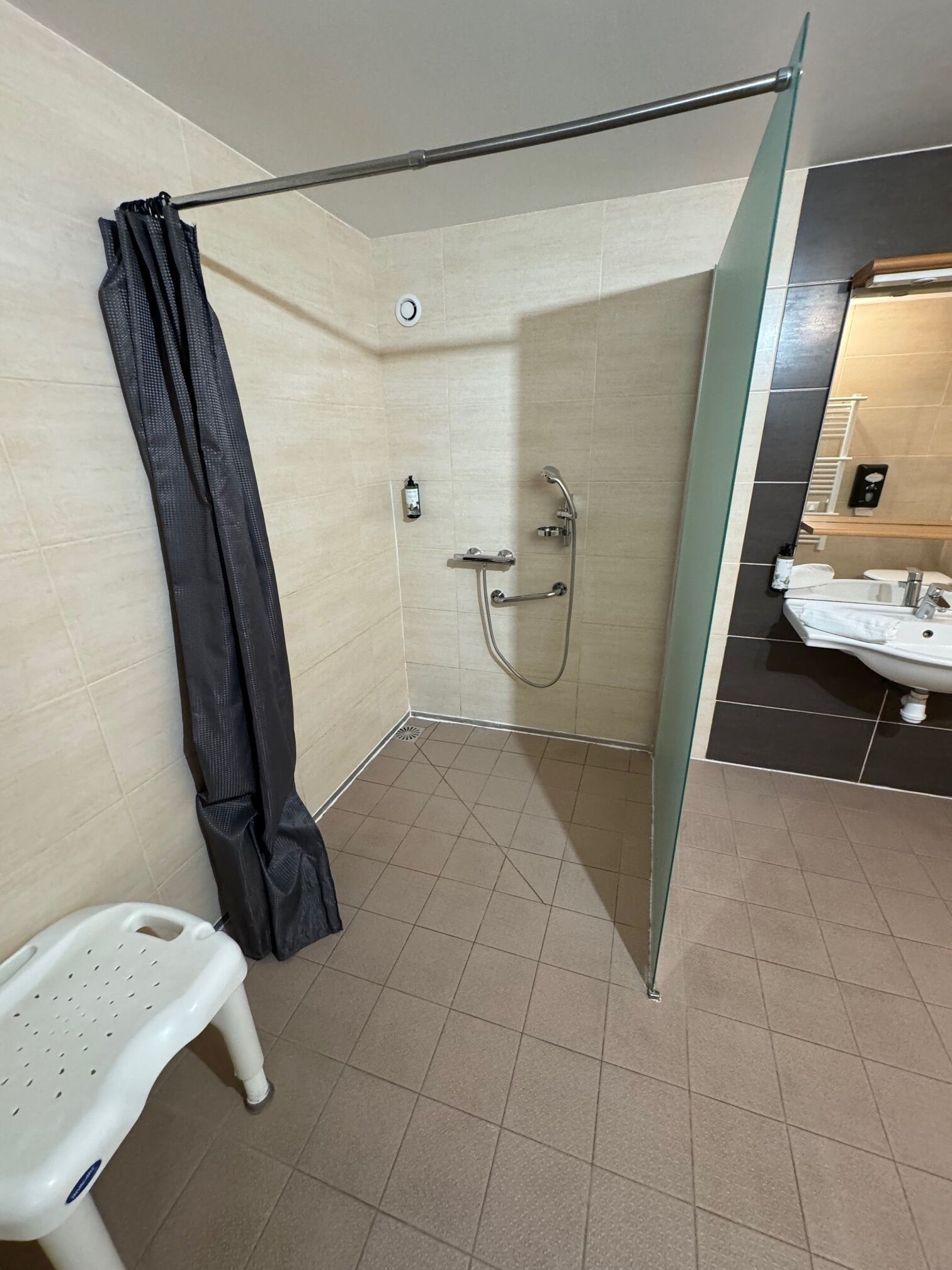 Ducha con puerta de vidrio, un taburete de plástico blanco, y un baño con lavamanos y espejo.