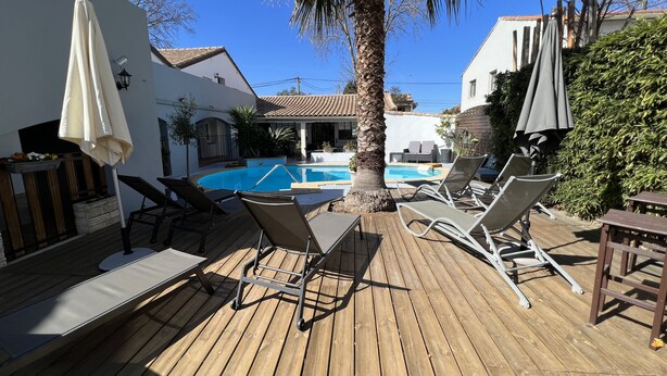 A wooden deck with lounge chairs and umbrellas, a swimming pool, and a white building with a tiled roof. - Hotel le Médieval (Aigues-Mortes)