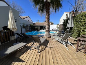 Una terraza de madera con sillones de descanso y sombrillas, una piscina y un edificio blanco con techo de tejas.