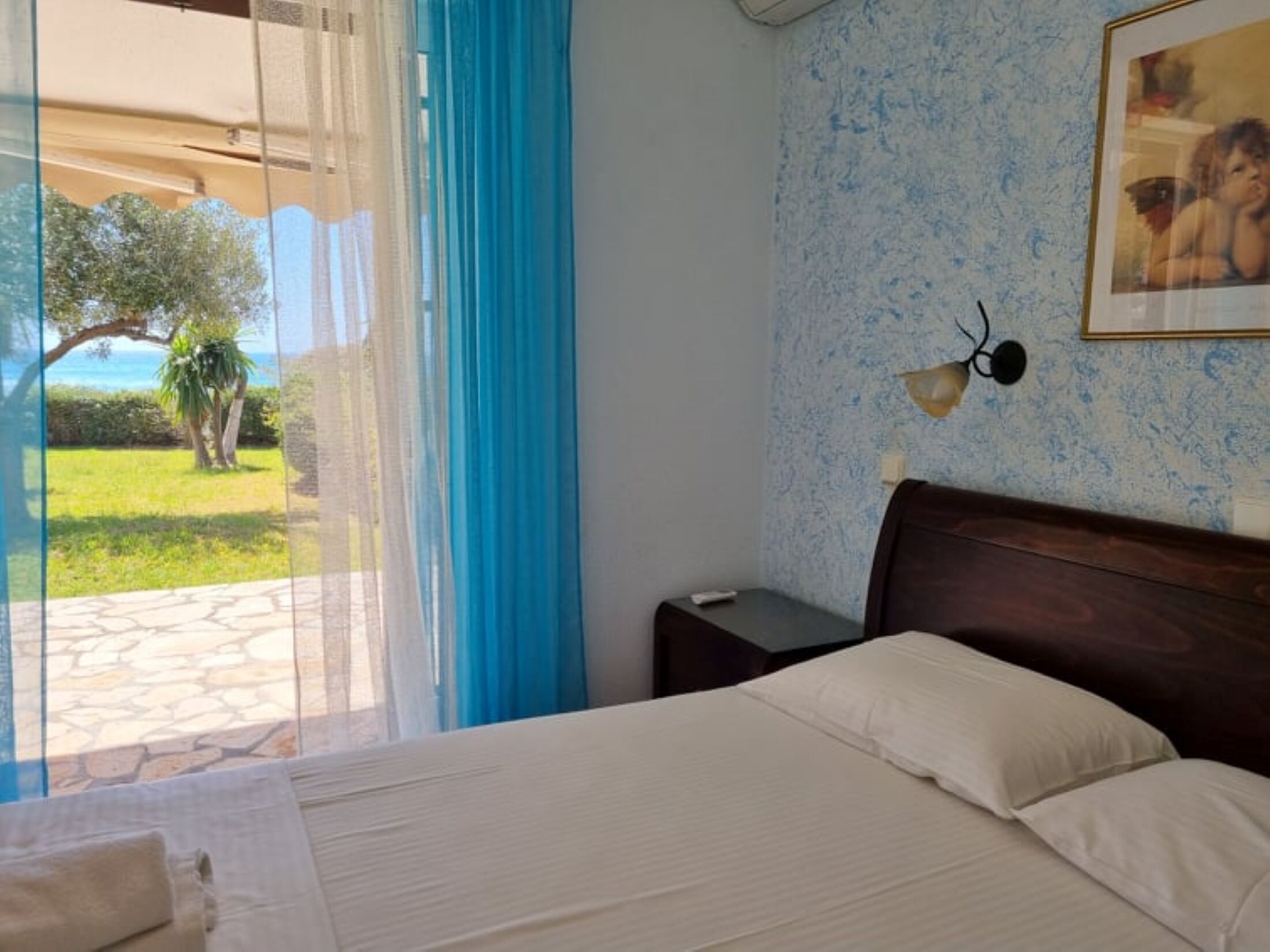 A bedroom with a bed, a nightstand, a lamp, and a view of a garden and sea through a window.