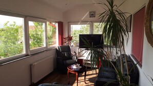 Ein Wohnzimmer mit einer schwarzen Ledersofa, einem Holztisch, einem Fernseher auf einem Schrank und einem großen Fenster mit Blick auf Bäume und Gebäude.