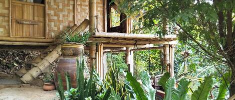Una estructura rústica de bambú con techo de paja, rodeada de abundante vegetación y plantas en macetas.