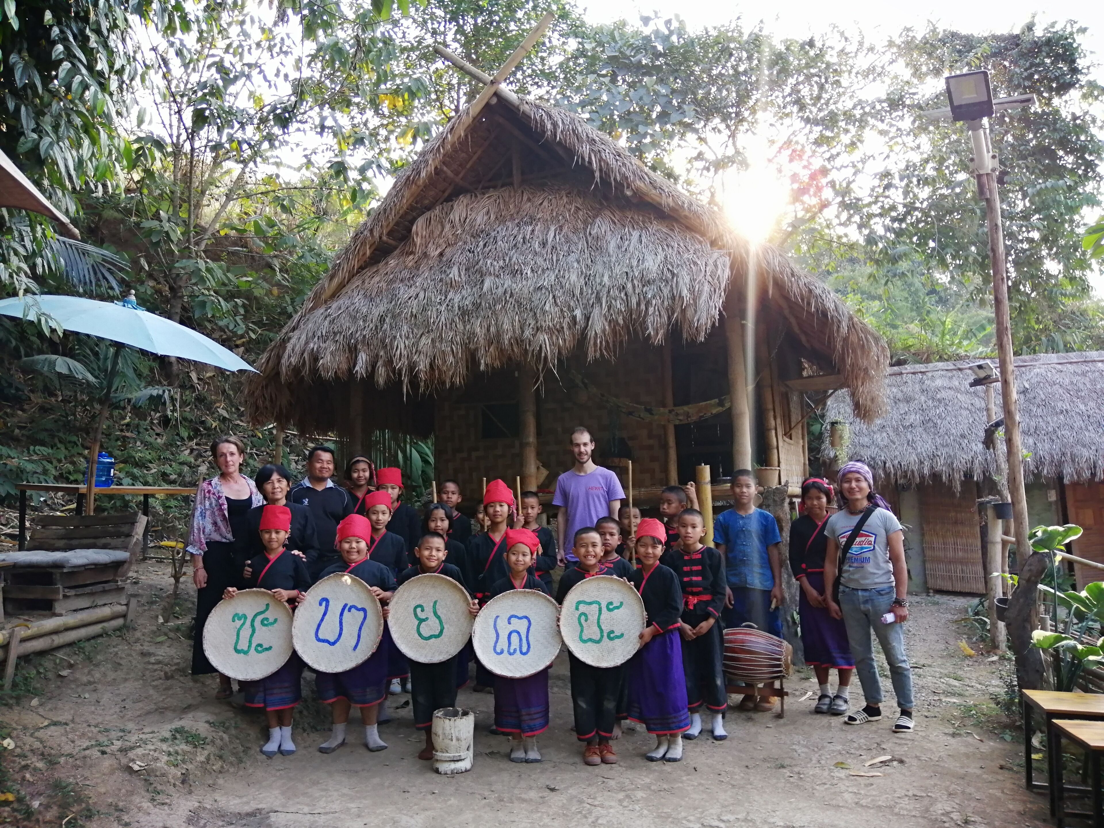 A group of people standing in front of a thatched-roof hut with a camera on a tripod nearby.