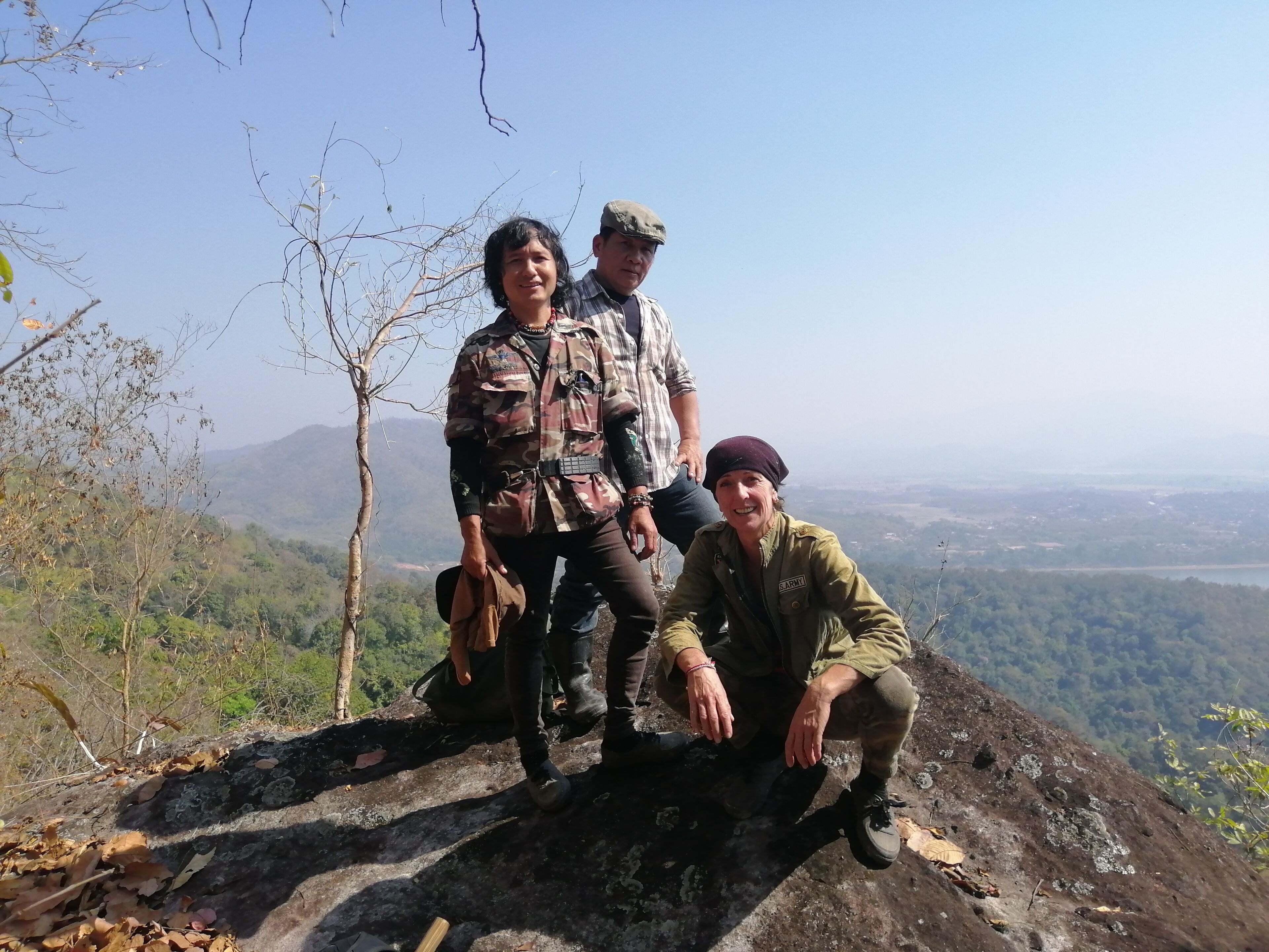 Three people on a rocky outcrop with a view of a valley and mountains in the background.