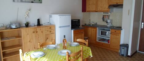 Una cocina compacta con armarios de madera, una mesa de comedor lista para cuatro personas, un refrigerador, un microondas y una lámpara colgante.