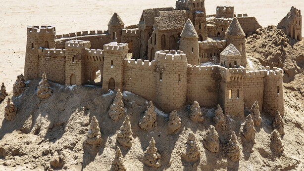 A sandcastle with multiple towers and walls, located on a beach with people and umbrellas in the background.