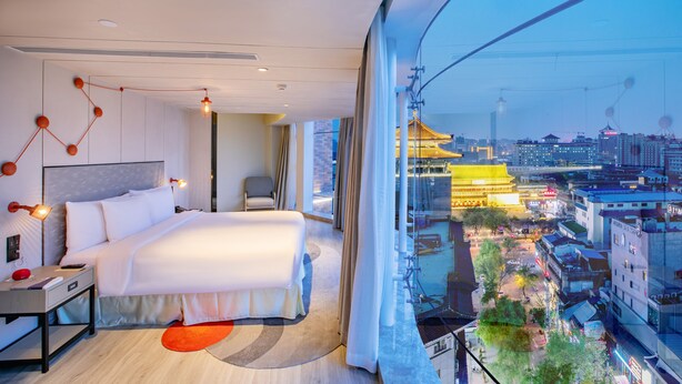 Une chambre d’hôtel moderne dotée d’un grand lit, d’un bureau et offrant une vue sur le paysage urbain grâce à une grande fenêtre.