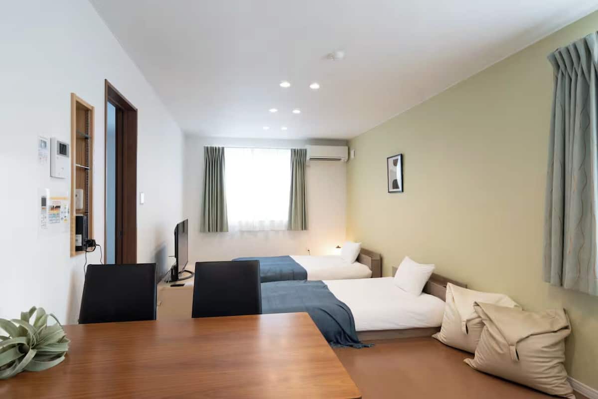 Une chambre d’hôtel avec une table à manger en bois, deux lits simples, une télévision et une fenêtre avec des rideaux.