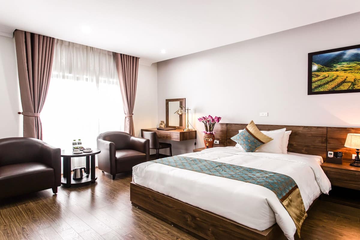 A hotel room with a large bed, two armchairs, a small table with a kettle, a bedside table with a lamp, and a painting of a landscape on the wall.