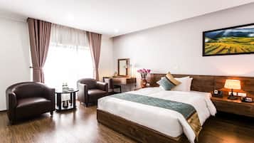 A hotel room with a large bed, two armchairs, a small table with a kettle, a bedside table with a lamp, and a painting of a landscape on the wall.