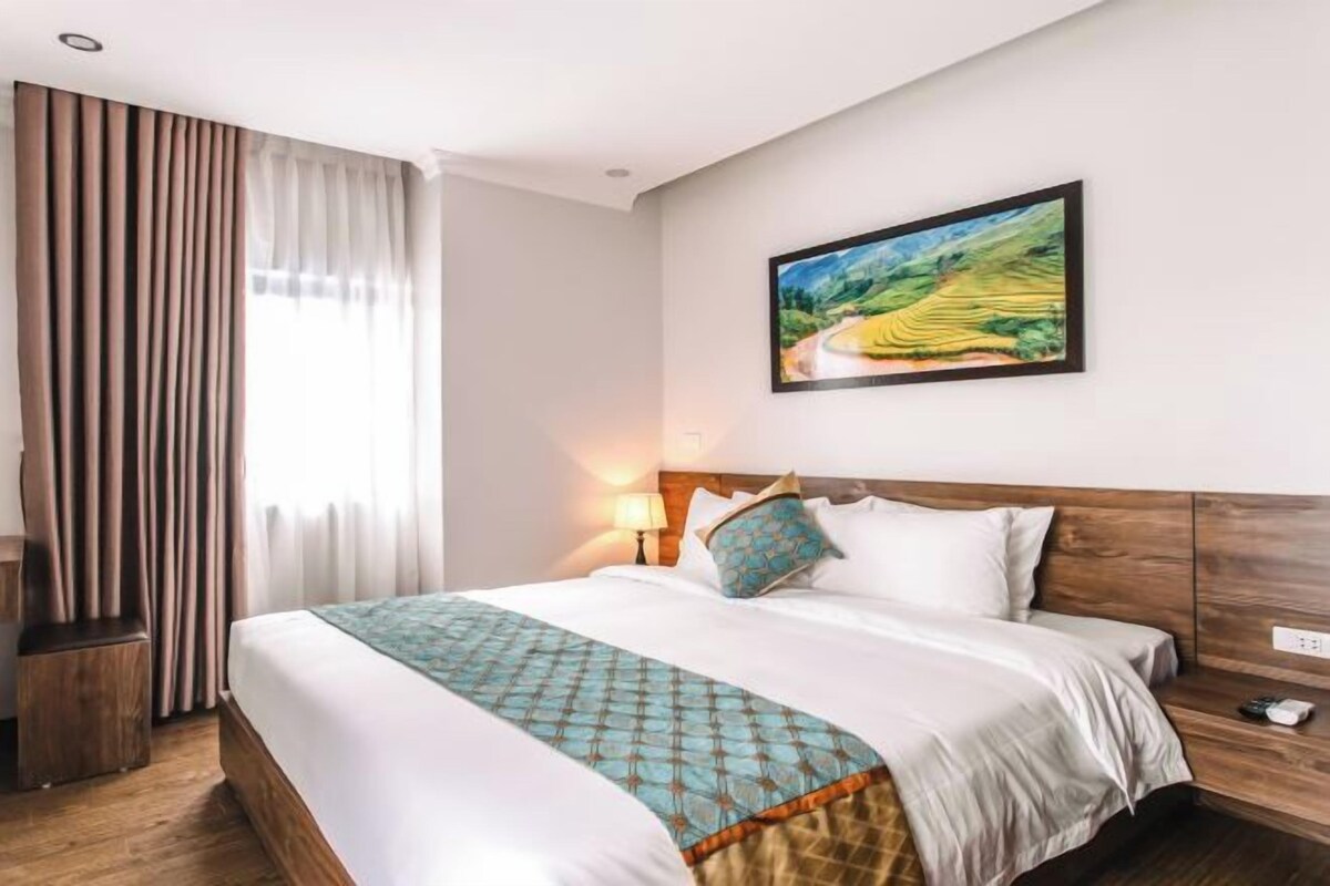 A hotel room with a large bed, a nightstand, a lamp, and a painting of a landscape on the wall.