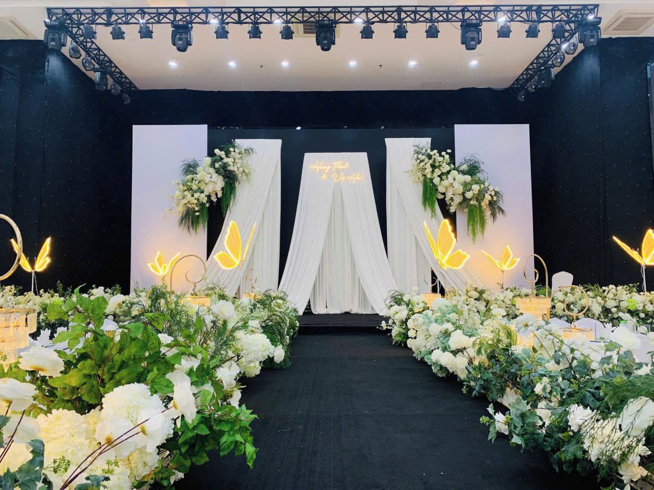 A stage with a black backdrop, white drapery, and floral arrangements.