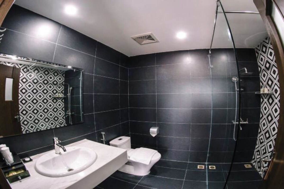 Une salle de bain moderne dotée d’un grand lavabo, de toilettes et d’un espace douche, avec des carreaux foncés et un motif géométrique sur les murs.