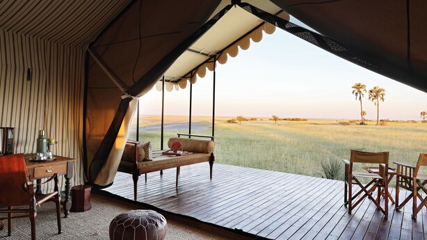 A tent with a wooden deck, a table, chairs, and a view of a grassy field with palm trees. - San Camp (Central Kalahari Game Reserve)