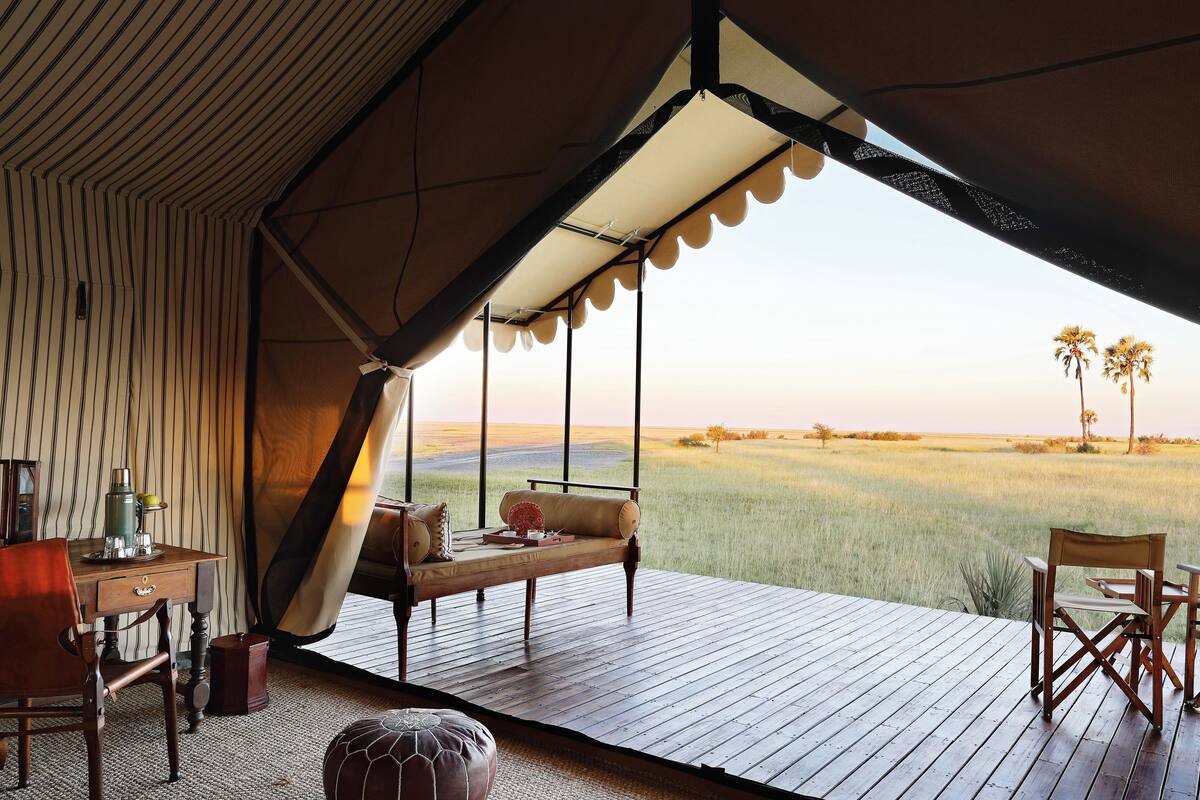A tent with a wooden deck, a table, chairs, and a view of a grassy field with palm trees.