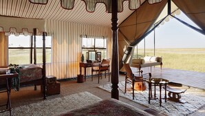 A tented room with a bed, a desk, and chairs, featuring striped curtains and a view of the landscape. - San Camp (Central Kalahari Game Reserve)