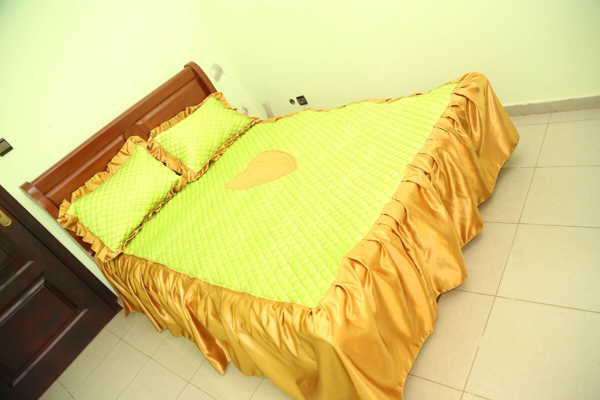 A bed with a yellow quilt and orange ruffled edges, a wooden headboard, and a plain wall with a light switch.