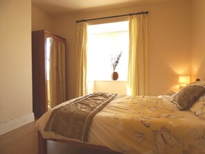 Un dormitorio con cama, una ventana con cortinas, una lámpara de noche y un espejo.