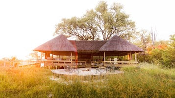Situated in a serene natural setting, this pavilion features a central fire pit surrounded by seating areas and a wooden deck.