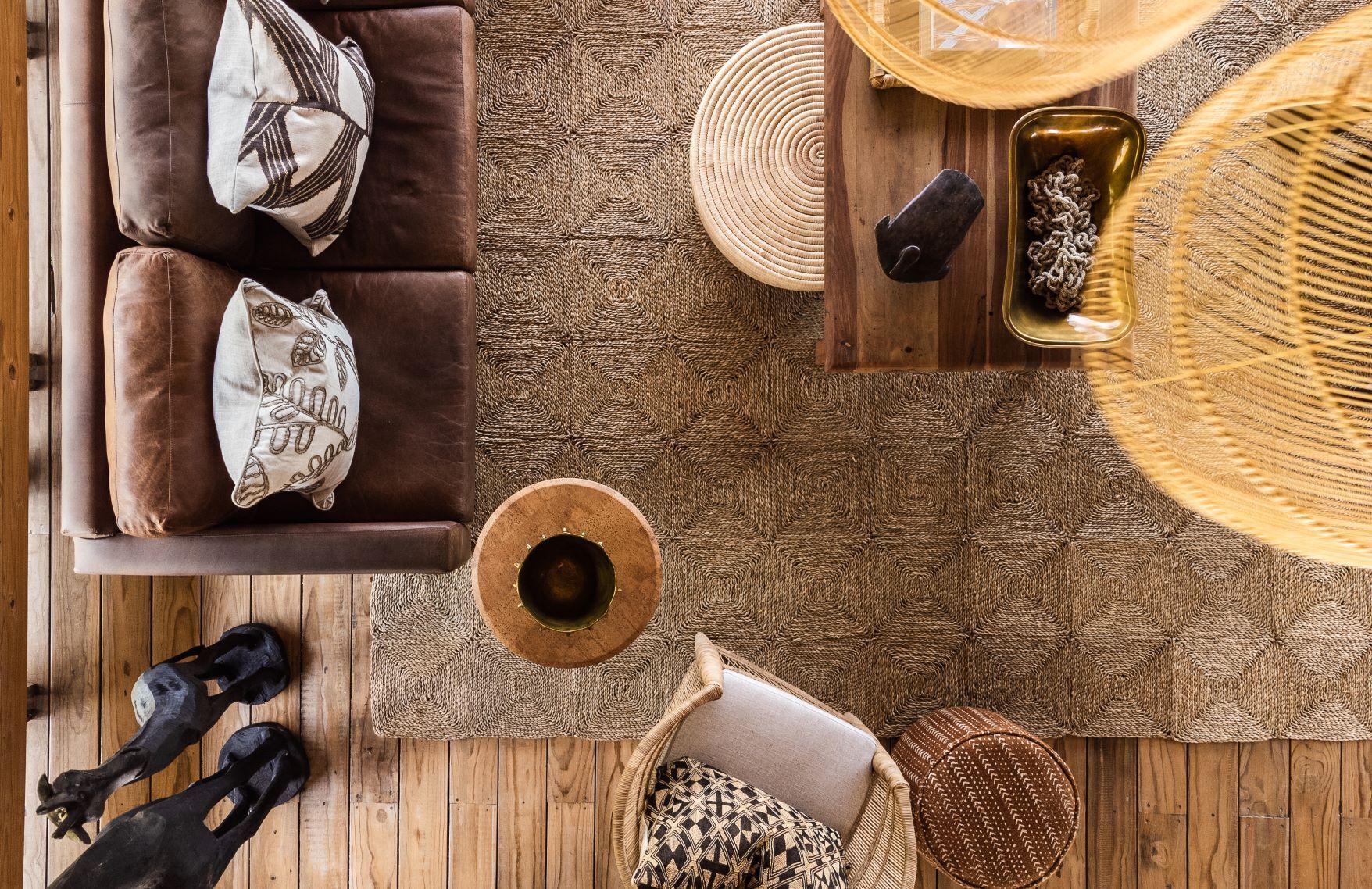 A top-down view of a cozy living room with a brown leather sofa, patterned cushions, a woven rug, a wooden coffee table, a woven basket, and a wicker chair.