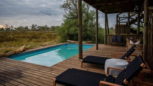 Una terraza de madera con piscina, sillas de descanso y un área cubierta con mobiliario de mimbre.