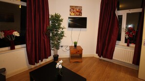 A room with a TV mounted on the wall, a wooden nightstand with a plant, and a window with red curtains. - PearTree House (Derby)