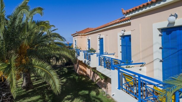 A row of white buildings with blue shutters and balconies, surrounded by palm trees. - Casa Kounopetra (Kefalonia)