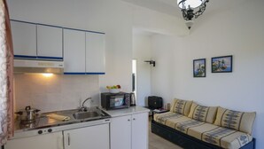 A compact kitchen with white cabinets, a stove, and a microwave. There is a sofa with patterned cushions and a small table with a speaker and a book. - Casa Kounopetra (Kefalonia)