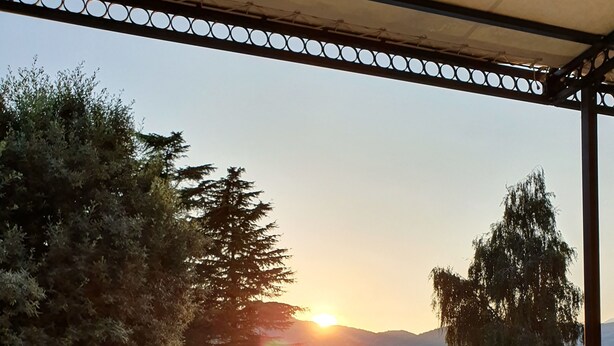 A dining table set with a menu, wine glass, and cutlery on an outdoor terrace with a pool and lounge chairs, overlooking a sunset and trees.