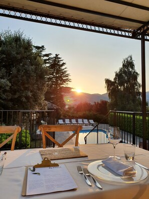 A dining table set with a menu, wine glass, and cutlery on an outdoor terrace with a pool and lounge chairs, overlooking a sunset and trees. - Agriturismo di charme Erbadoro (Alvito)