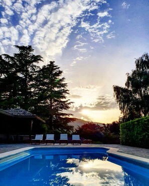 Piscina con sillas de descanso y un pabellón, rodeada de árboles y vistas al atardecer.