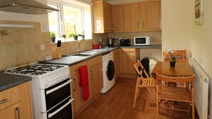 A compact kitchen with a gas stove, oven, microwave, and sink. There is a wooden dining table with chairs and a window with plants. - Cowsley House (Derby)