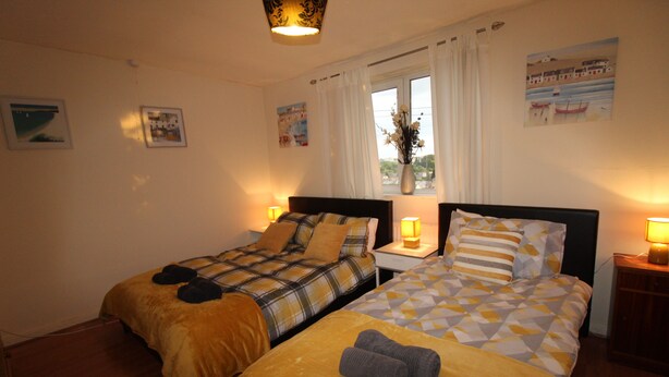 A hotel room with two beds, a window with curtains, a vase with flowers, and wall art. - Cowsley House (Derby)
