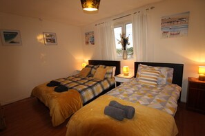 A hotel room with two beds, a window with curtains, a vase with flowers, and wall art. - Cowsley House (Derby)
