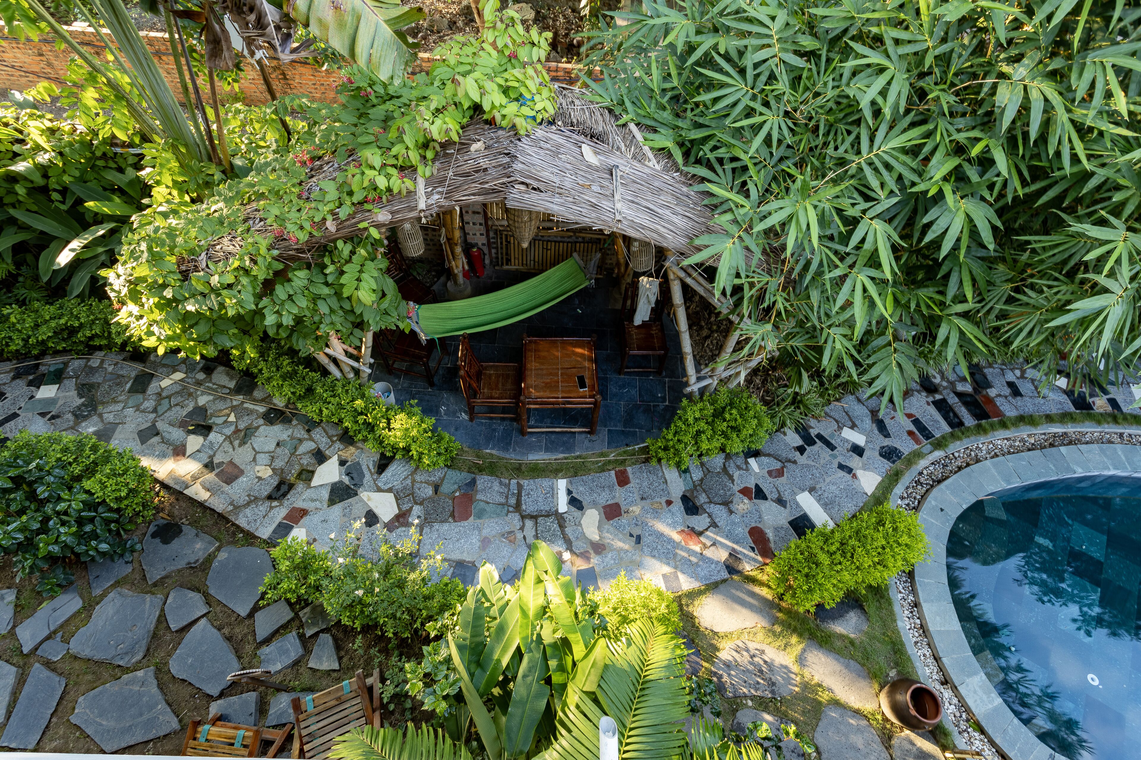 Una cabaña con techo de paja y una hamaca, rodeada de abundante vegetación y un área empedrada que conduce a una piscina.