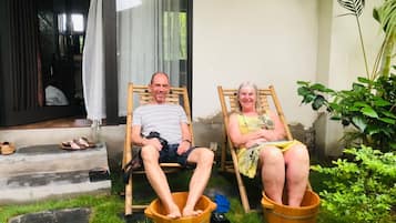 Dos personas sentadas en cubos de madera al aire libre, con la entrada de una casa y follaje de fondo.