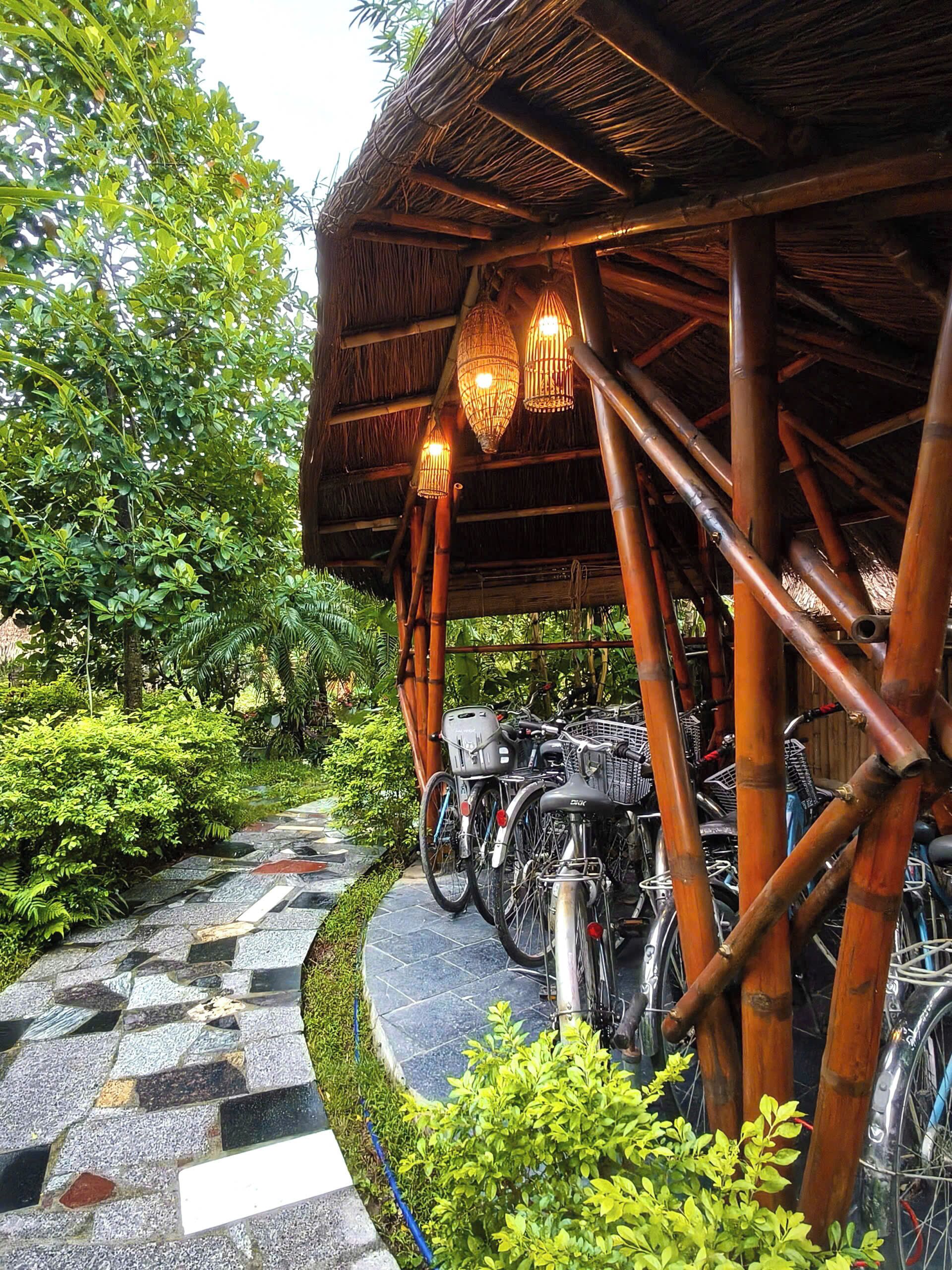 Un espace extérieur abrité où sont garés des vélos, sous une structure en bambou, le tout entouré d’une végétation luxuriante et bordé d’un sentier en pierre.