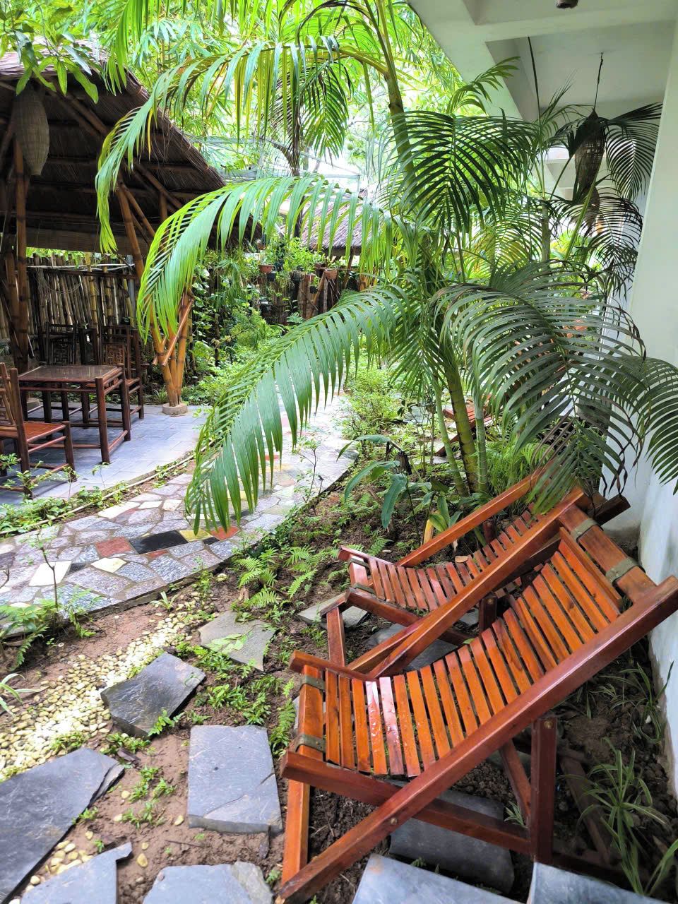 Una silla de madera para lounge en un jardín tropical con senderos de piedra y abundante vegetación.