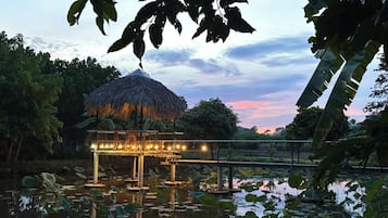 A thatched-roof structure with a railing, surrounded by water lilies and trees.