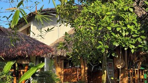 Un sendero conduce a una casa tradicional con techo de paja, rodeada de abundante vegetación y bambú.
