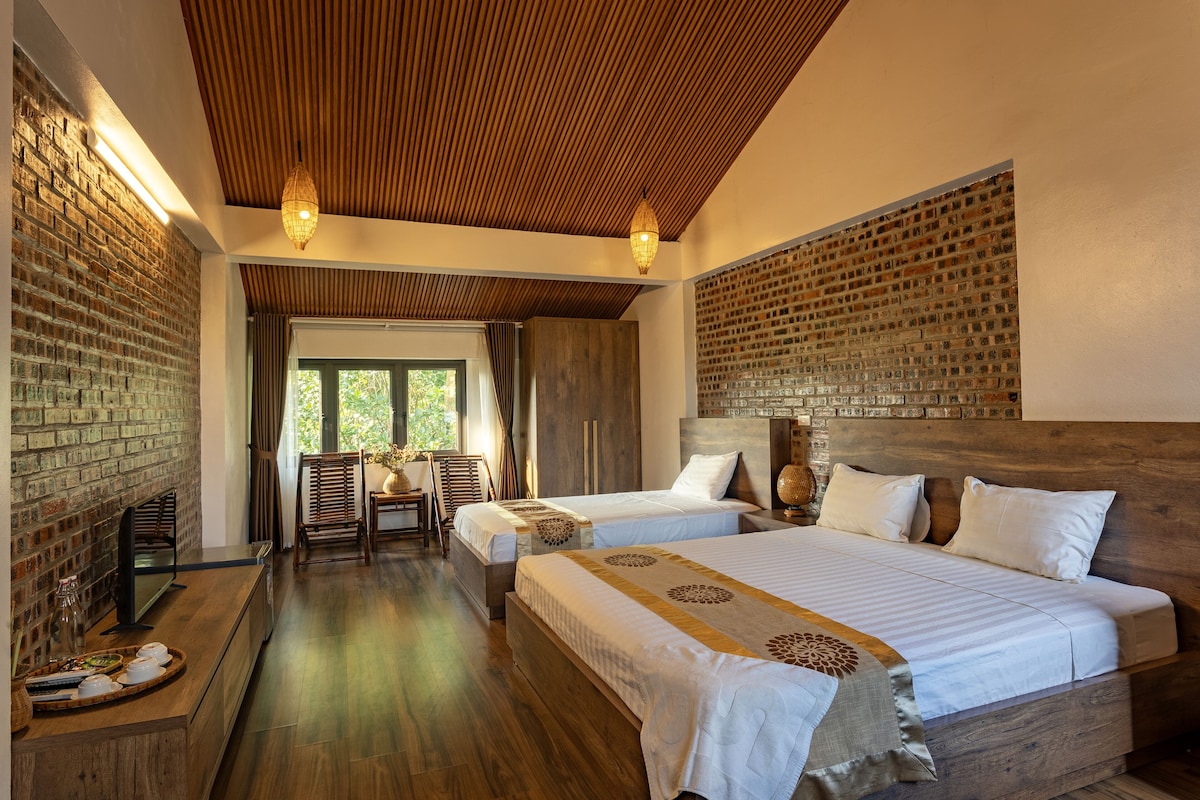 Quarto de hotel com duas camas, teto de madeira, paredes de tijolo e vista para o verde lá fora.