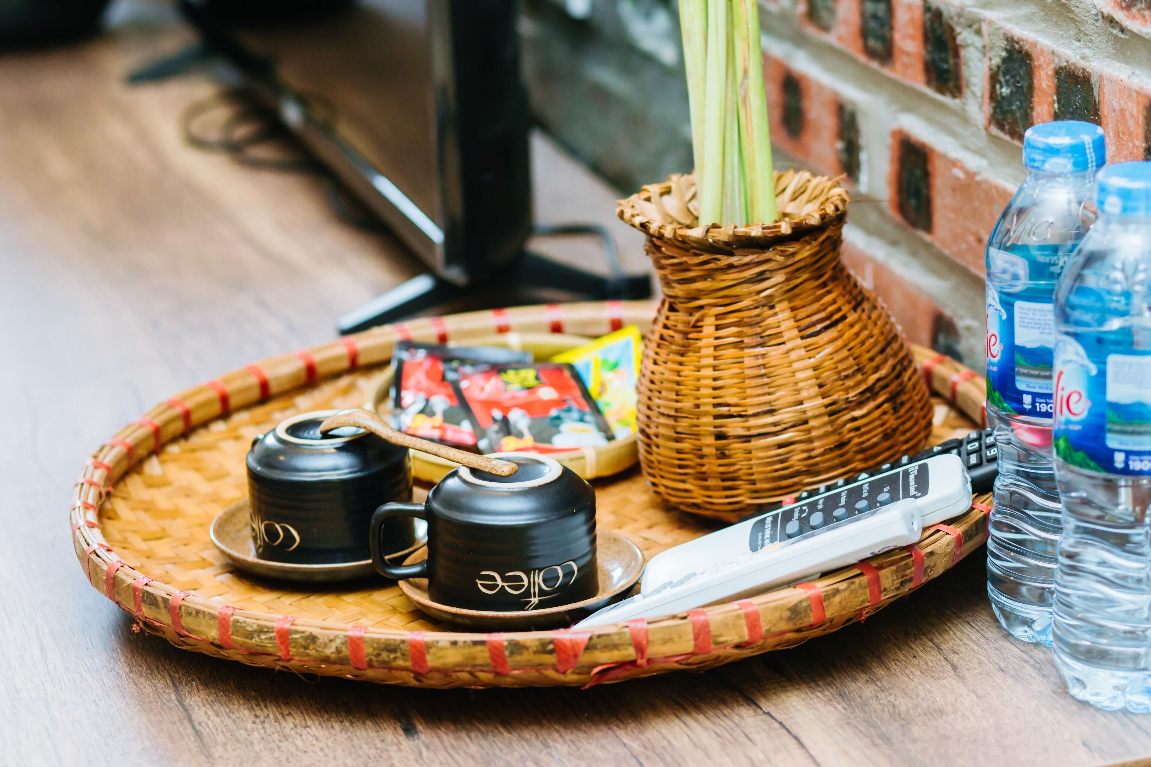 A tray with a woven basket containing a teapot and cups, a remote control, and two water bottles.